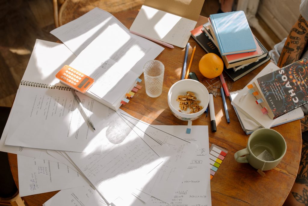 Papers and books sprawled out on a table with a mug and bowl pf cereal, with streaks of sunlight coming in through a window