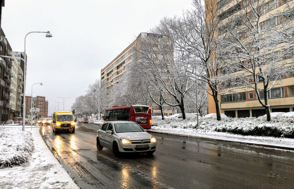 A slushy & snowy winter day. A road is pictured with cars and a bus, and grey/brown are the prominent colours. A lot of melted snow and sludge can be seen on the sidewalk and road