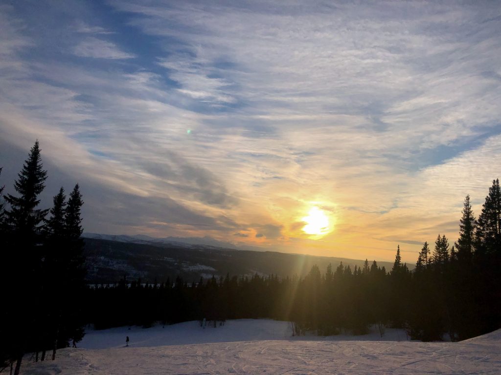 The view from a snowy mountaintop, trees on both sides of the slope. A sunset is pictured