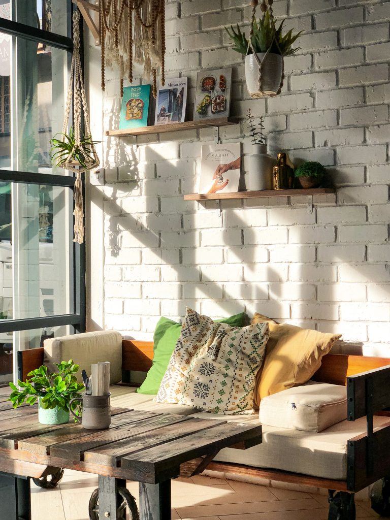 A cozy living roo witha. sofa and plants with sunlight streaming in through the window. A white brick wall lined with shelves is in the background
