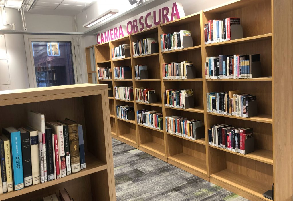 Warmly lit booskshelves, pink block letters say "CAMERA OBSCURA" atop them.