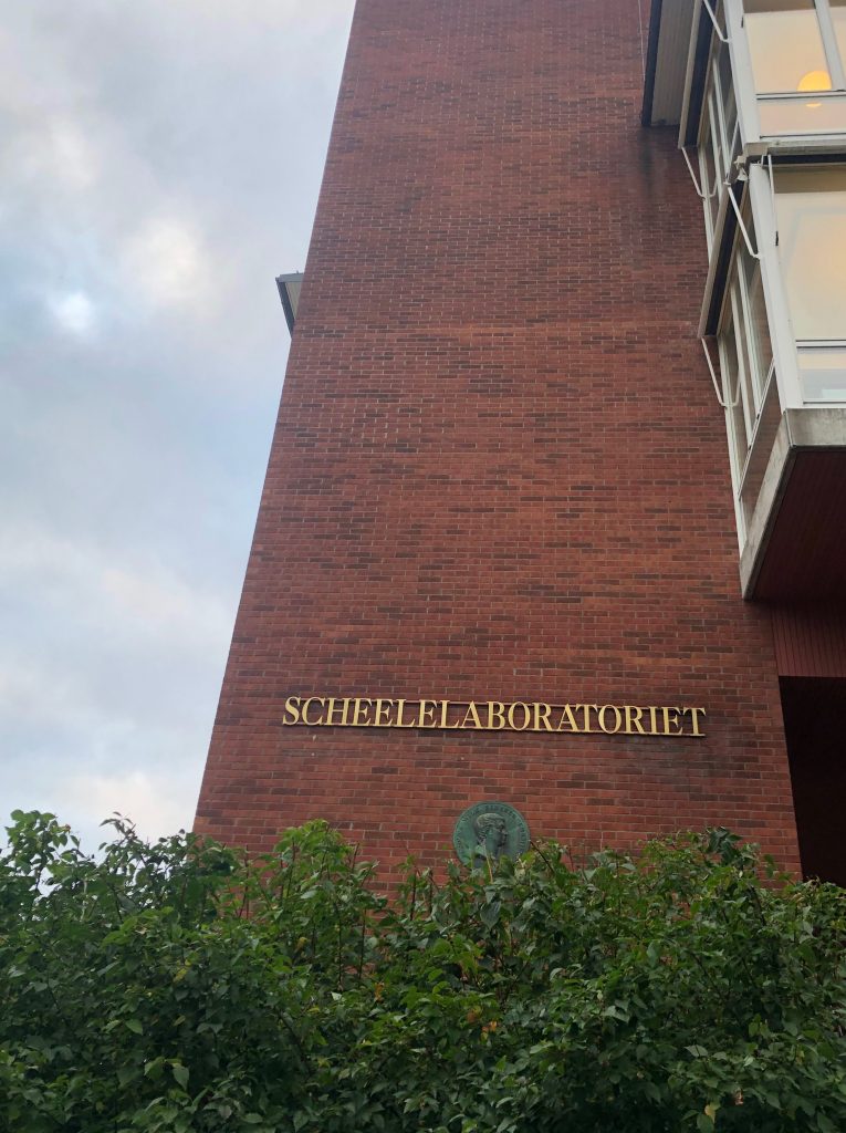 "SCHEELELABORATORIET" written on a red brick wall of the building, framed by bushes underneath.