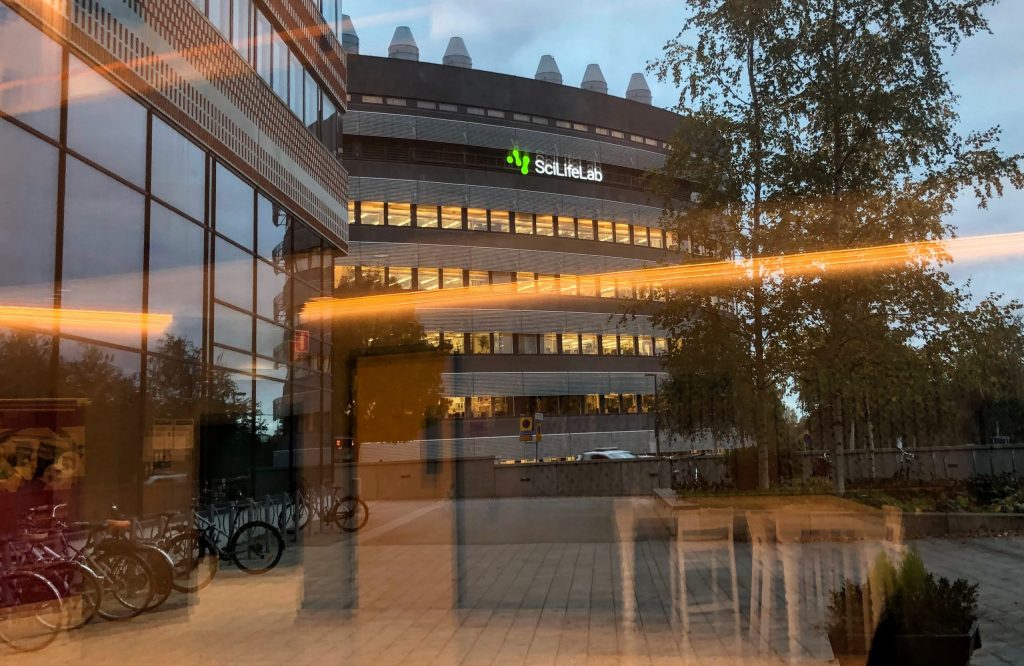 A grey building with "SciLifeLab" in green is seen through a large window on the ground floor. The inside of the building the photographer is in can be seen because there's a reflection in the glass.