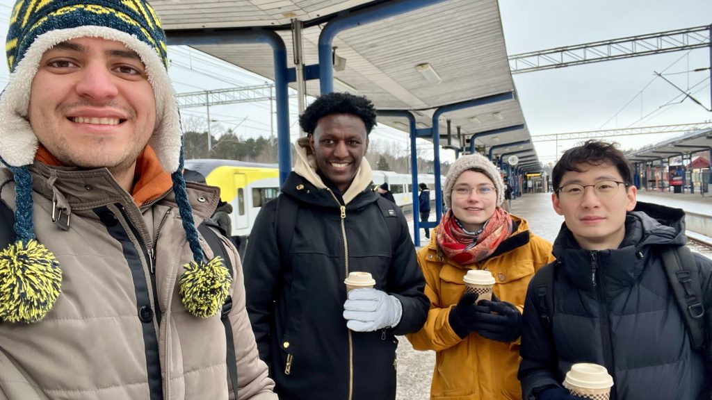 A selfie of four friends smiling and holding coffee cups at a train station on our trip to Uppsala