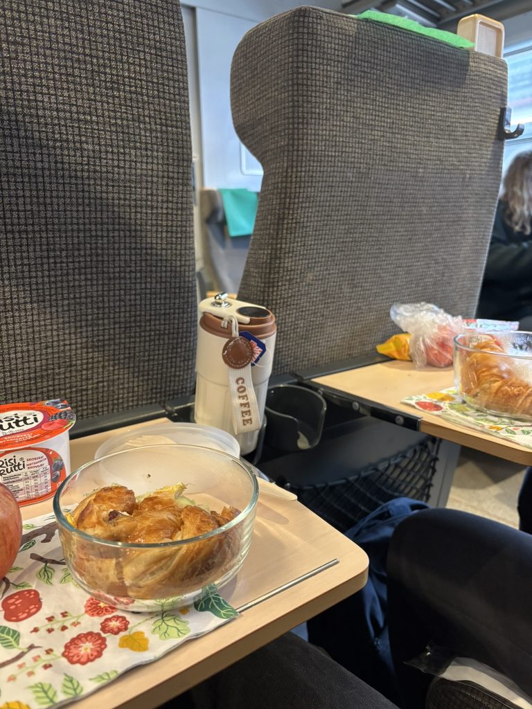 A shot of the table tray behind seats on trains, laden with breakfast items: a croissant sandwich, an apple, rice pudding and tea on the side. 