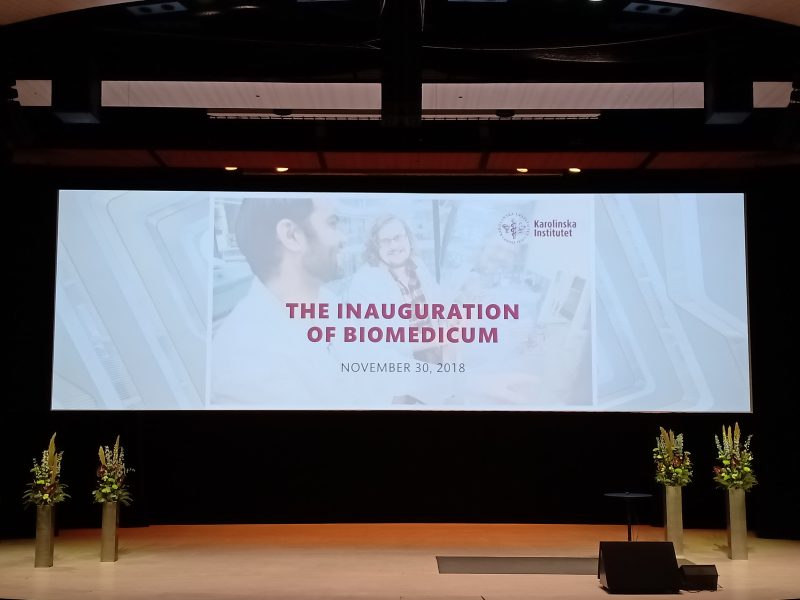 stage with a backdrop written with The Inauguration of Biomedicum November 30, 2018 and KI's logo; podium and decorative flowers