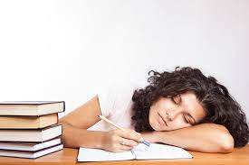 A tired female student with her head slumped down on the desk. She also has a pile of books beside her indicating she is tired and studying.