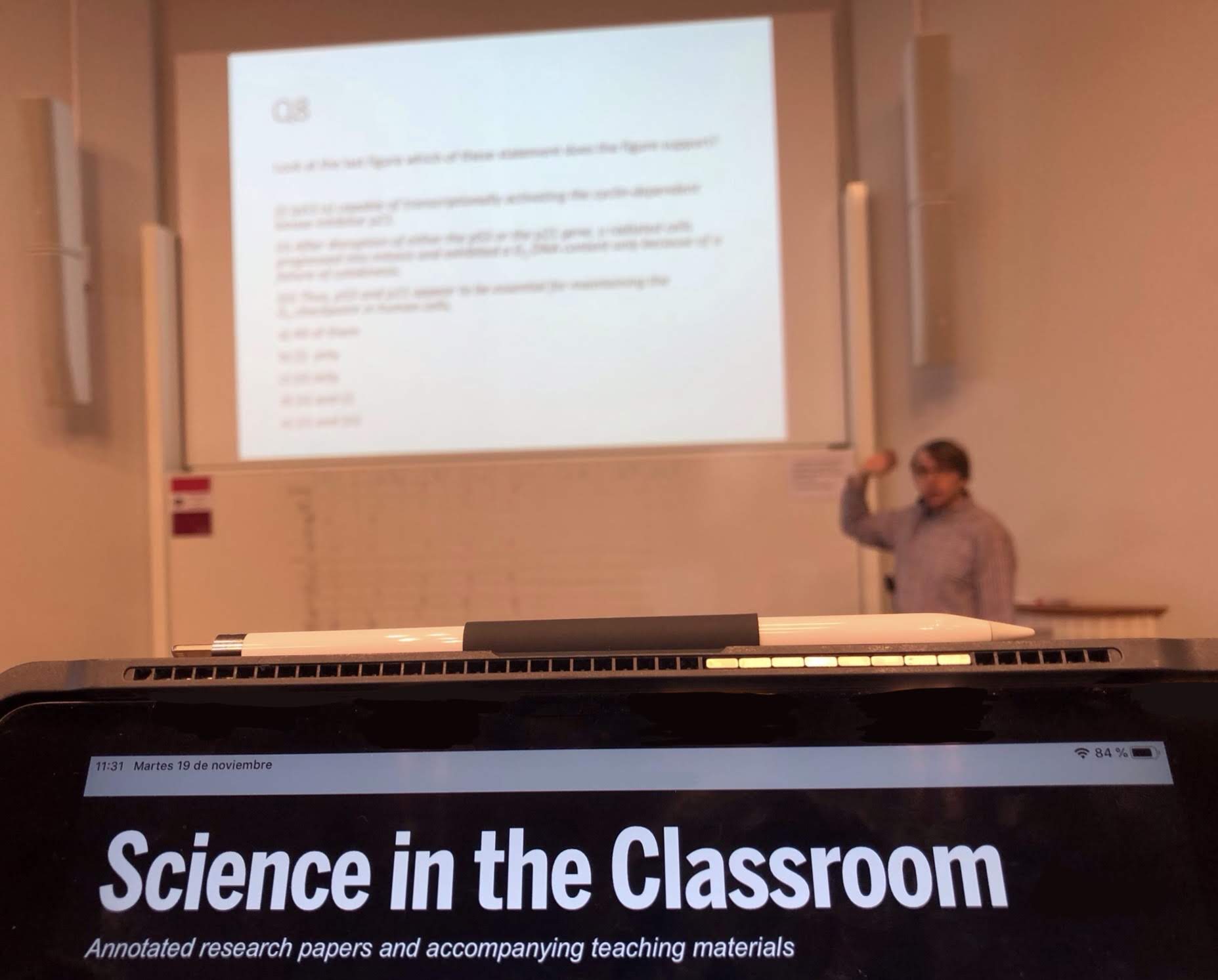 A teacher pointing to a projected screen in the background, with a device screen showing "Science in the Classroom" in the foreground. The subheading is "Annotated research papers and accompanying teaching materials
