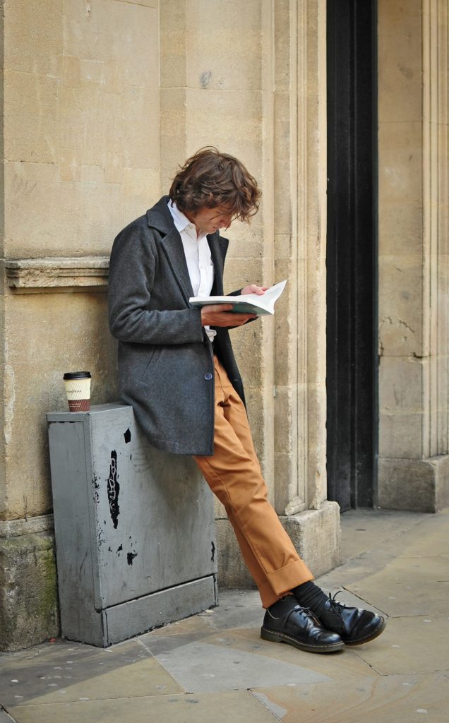 A curly haired man leaning against a metal box propped against a wall reading, coffee cup resting beside him.
