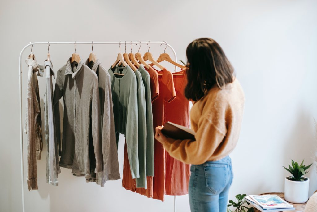 A person with shoulder length hair looks at a rack of clothes with their back towards the camera. The rack has shirts, tops, and t-shirts hanging. 