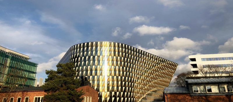 Aula Medica, a shiny building with a blue sky as backdrop. A green glassy Biomedicum rises on the left, and the Karolinska hospital on the right. A red brick building (MF Kårhuset) sits in front.