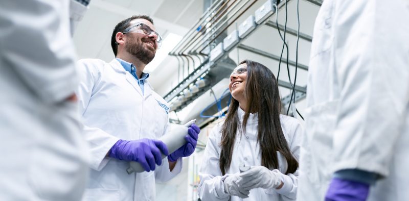 4 people in white coats stand in a circle, only 2 of them in view who are both wearing gloves and holding pipettes