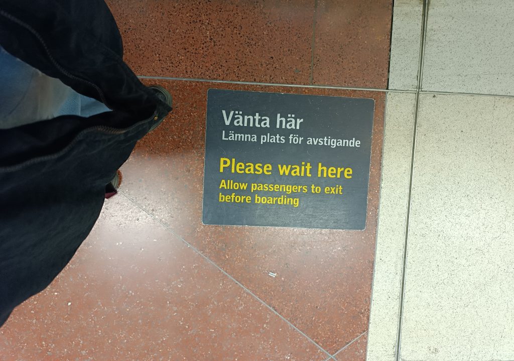 Sign on floor at train station saying "Please wait here. Allow passengers to exit before boarding", in Swedish and English.
