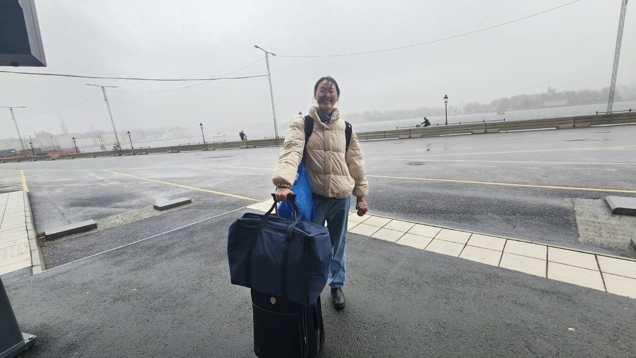 A girl standing with a cooler bag and a suitcase waiting for the bus in the gloomy cloudy weather.