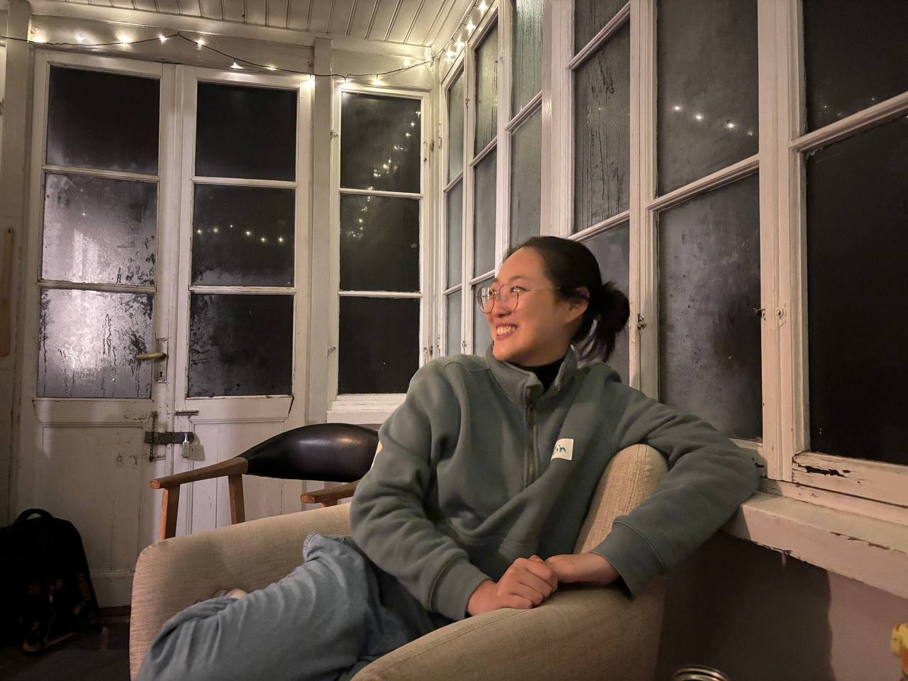 A girl sitting on the armchair looking to the side while smiling. The room is small and it's dark outside.
