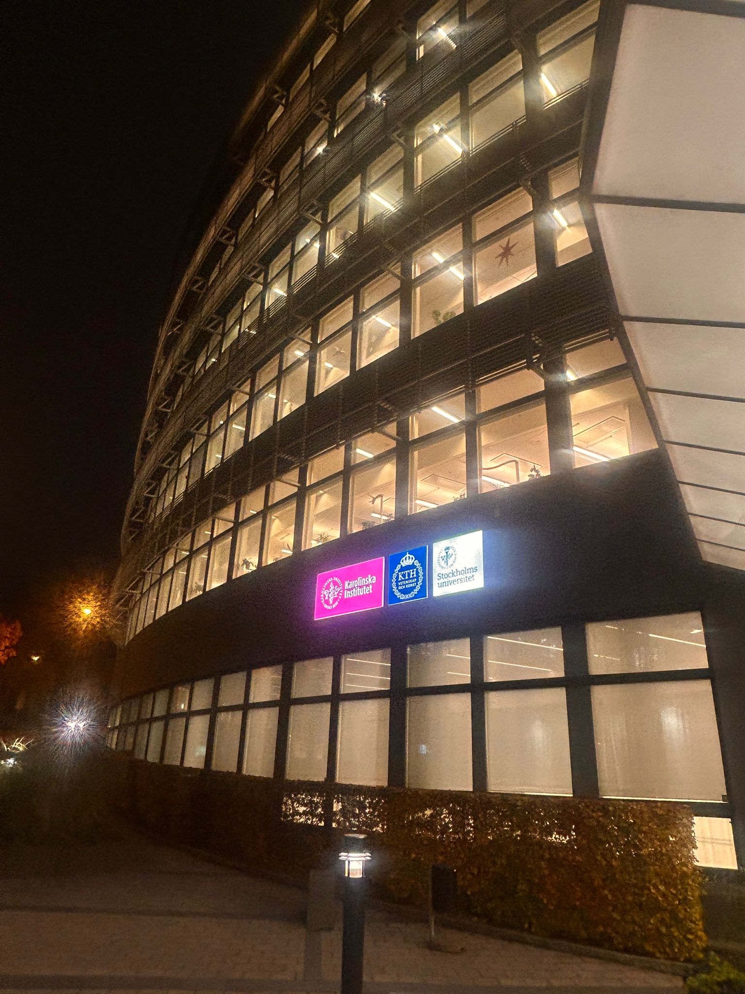 SciLifeLab building at night showing the logos of the three universities: Karolinska Institutet, KTH ans Stockholm University (SU).
