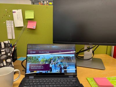 A desk set up with a laptop screen open, a desktop monitor, sticky notes, folder, cup of coffee and lunch bag