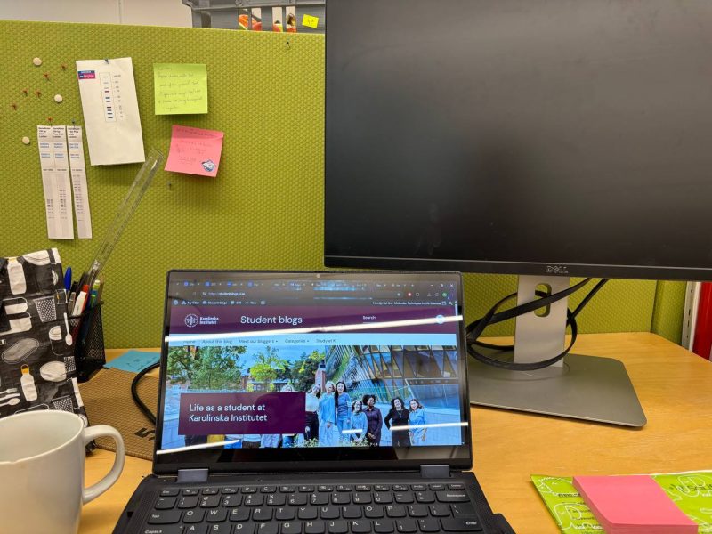 A desk set up with a laptop screen open, a desktop monitor, sticky notes, folder, cup of coffee and lunch bag
