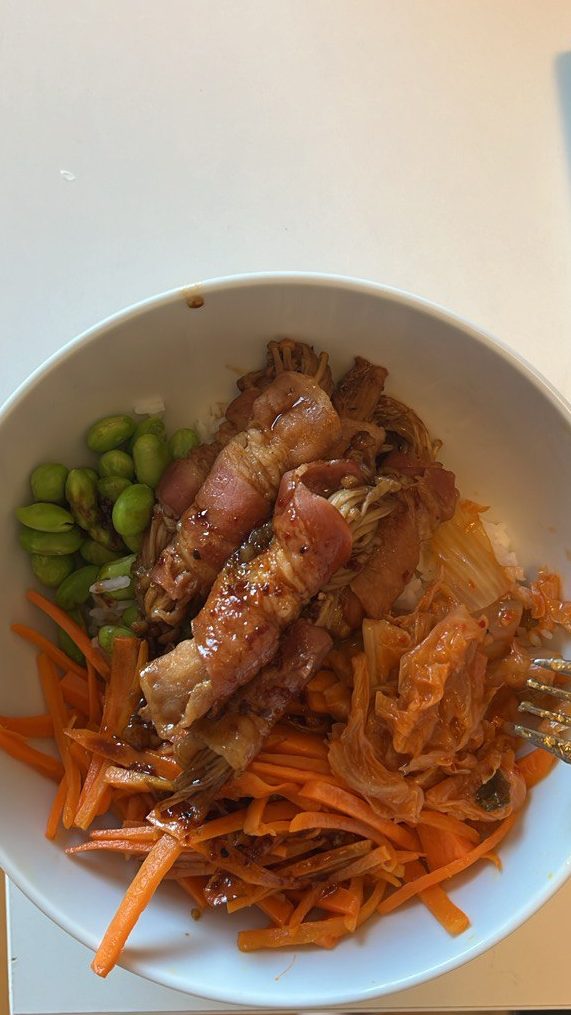 A bowl with edamame, shredded carrots, kimchi and bacon wrapped enoki mushrooms.