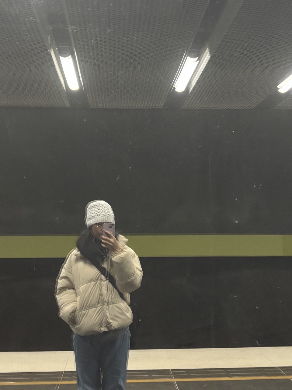A mirror selfie of a girl in winter clothes and hat in the subway station.