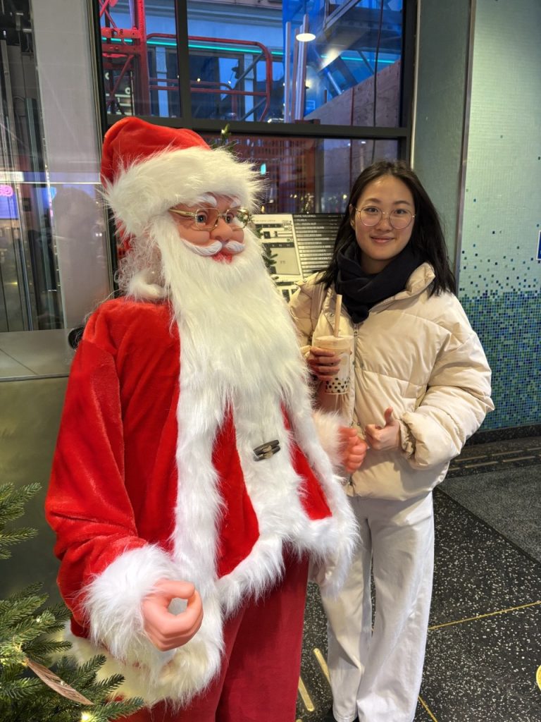 A decorative Santa Claus next to a girl dressed in winter clothes and holding a milk tea with boba.