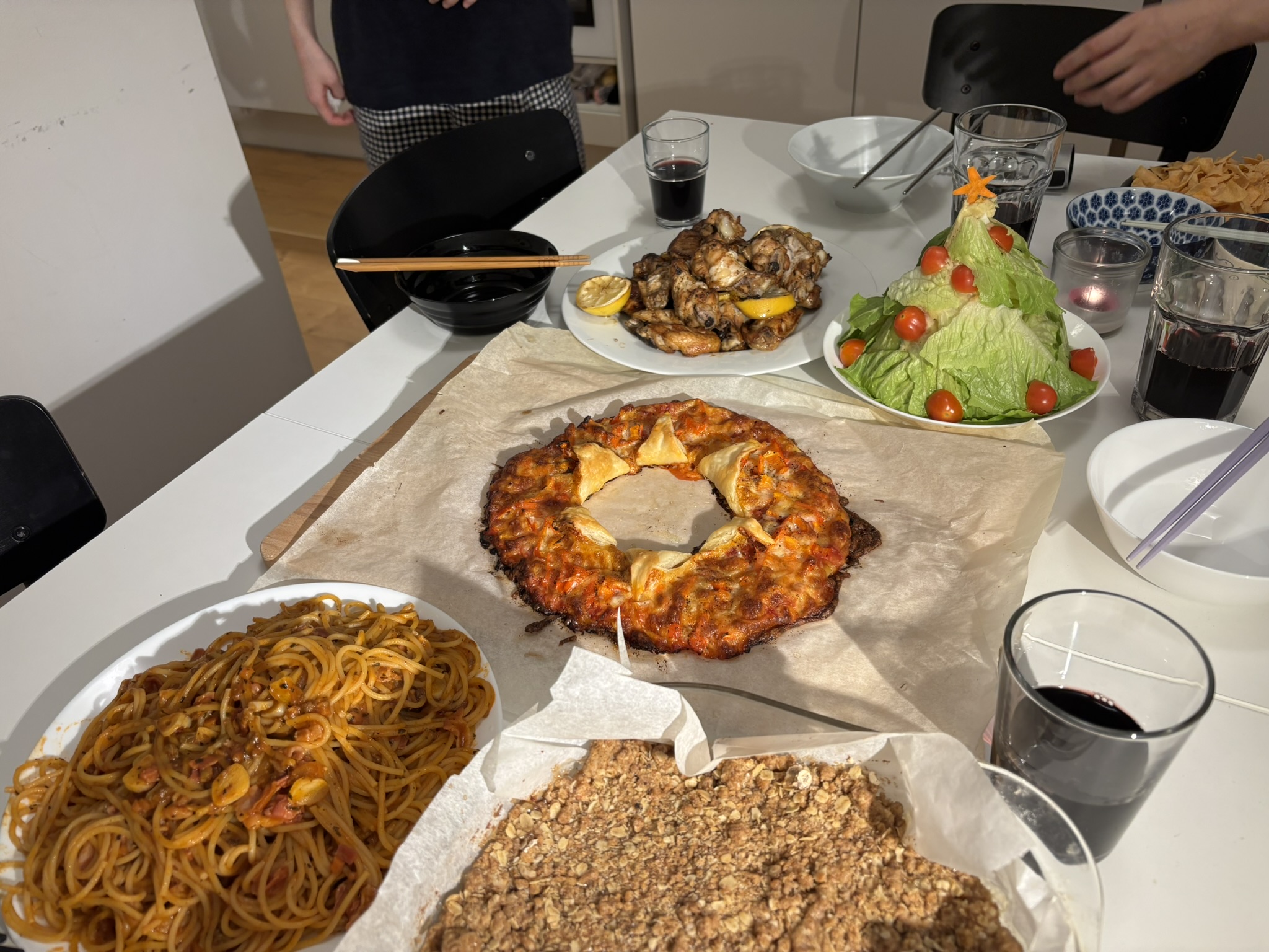 A table spread with pasta, apple crumble, a pizza baked wreath, an edible christmas tree made with lettuce and tomatoes, and some chicken wings.