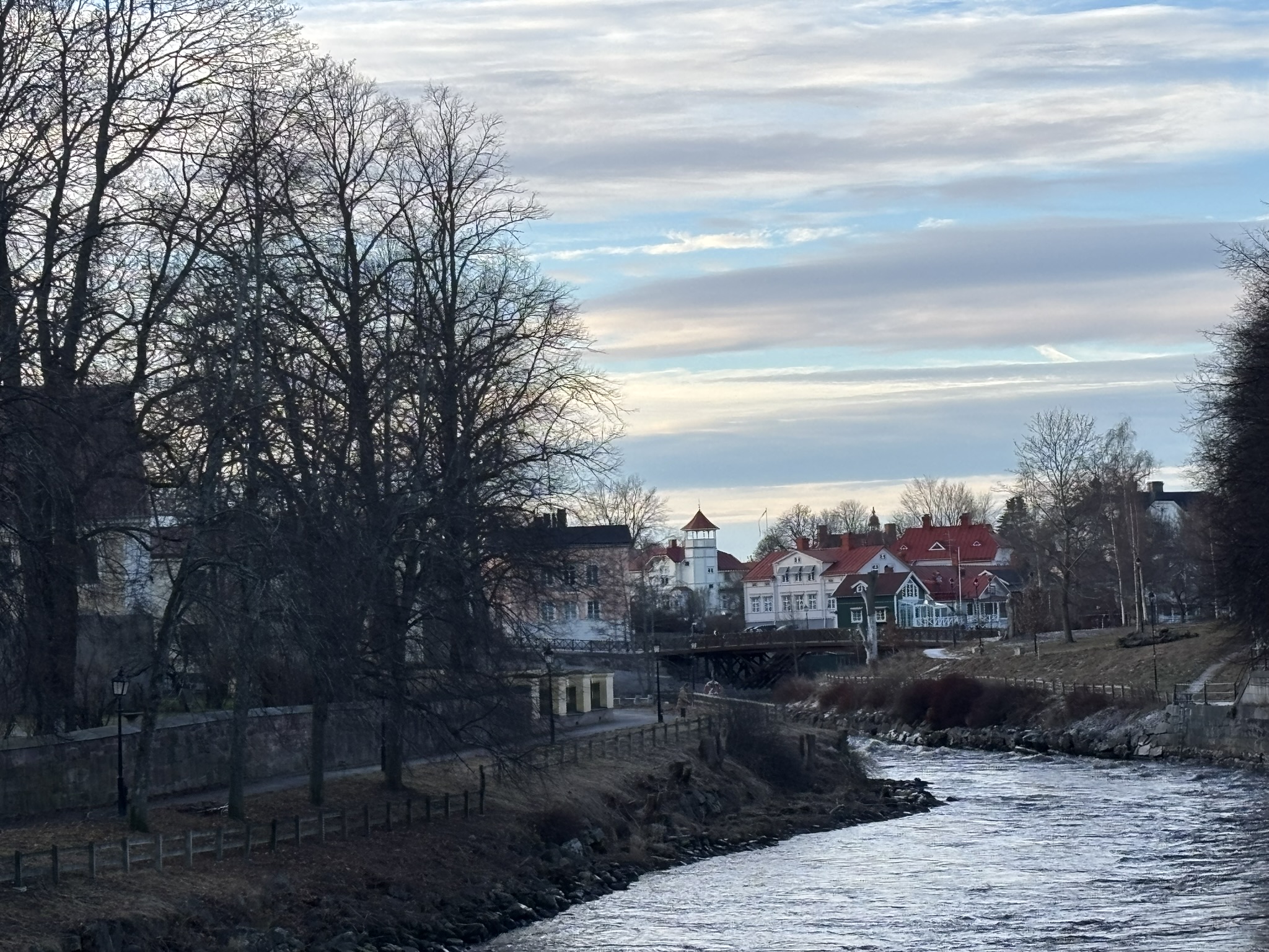 A wintery river flowing in the city lined with barren trees on the side. In the distance are houses topped with red roofs and white walls.