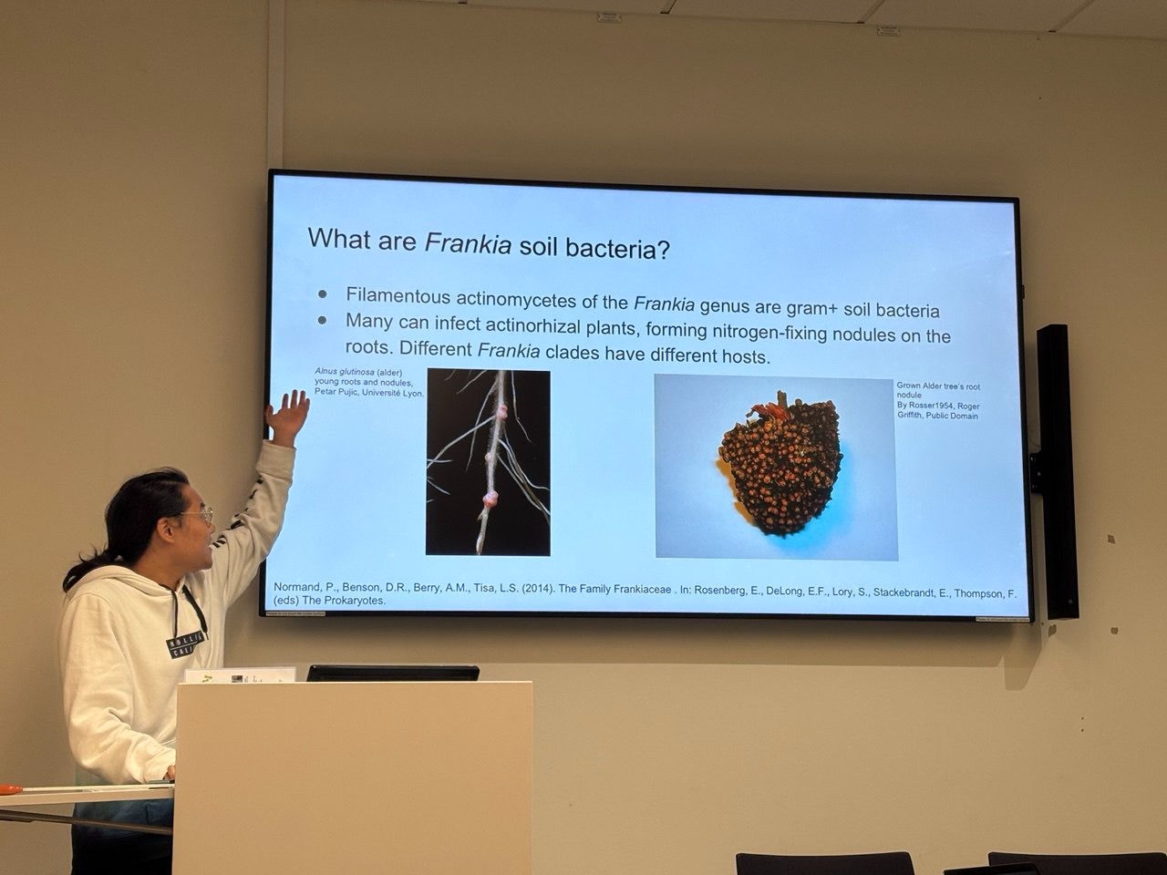 A male is presenting in front of a room of people (indicated by the back of two heads) and looking towards a large screen with material on his project.