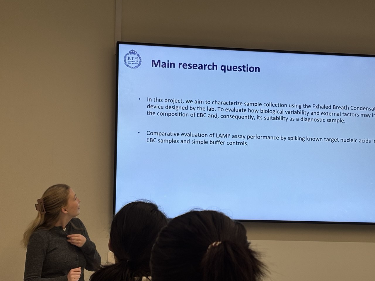 A female is presenting in front of a room of people (indicated by the back of two heads) and looking towards a large screen with material on her project.