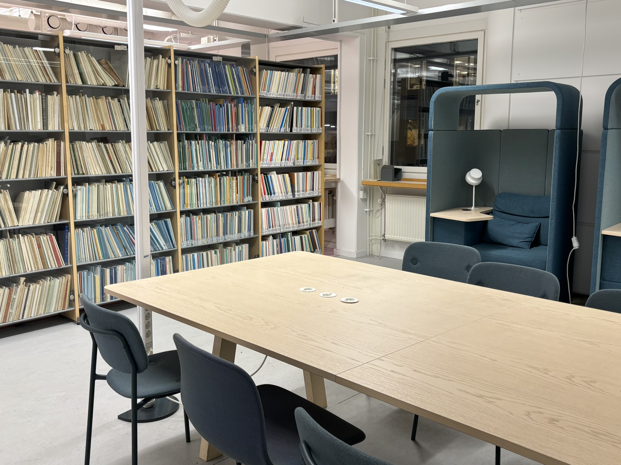 Background of shelves of books lined up; A long desk stretches out in front with chairs. A snapshot of the library