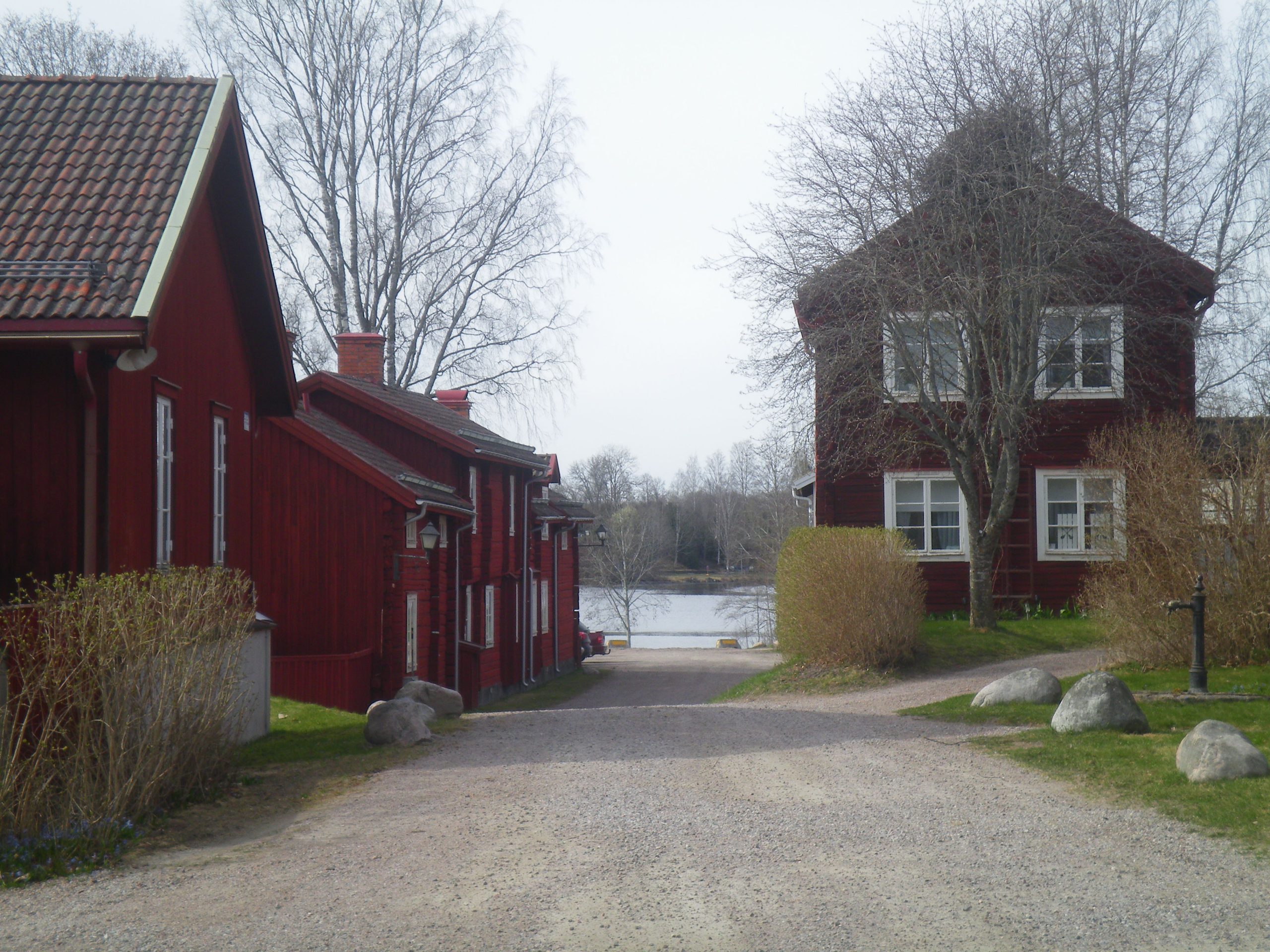 A dirt path with stones gflanked by red wooden houses leading to a farway lake seen in the background. 