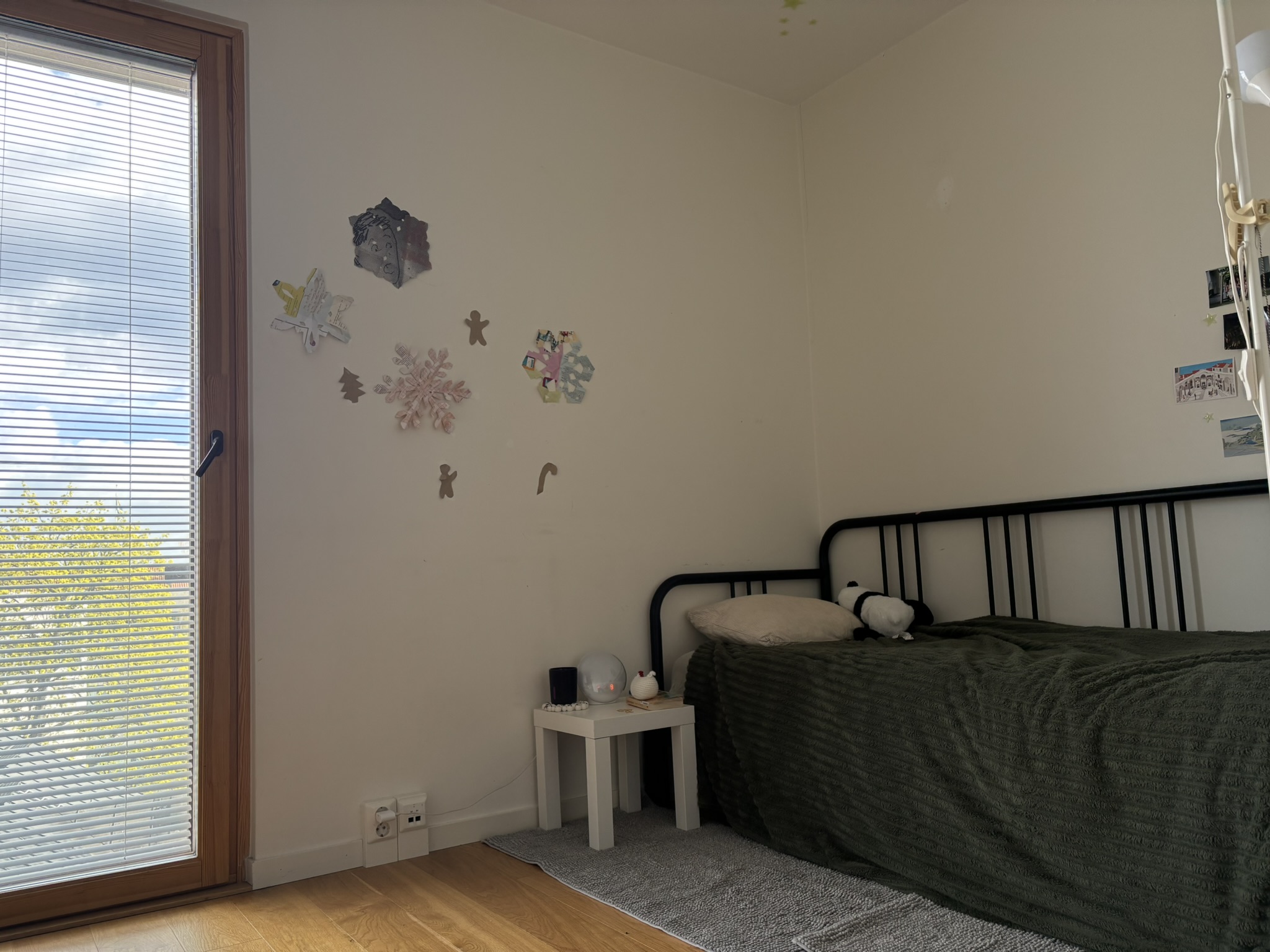 In the left corner of the room, there is a full length window showing the sunny day outside. On the right corner there is a single bed with green sheets, and a small side table to the left of it.