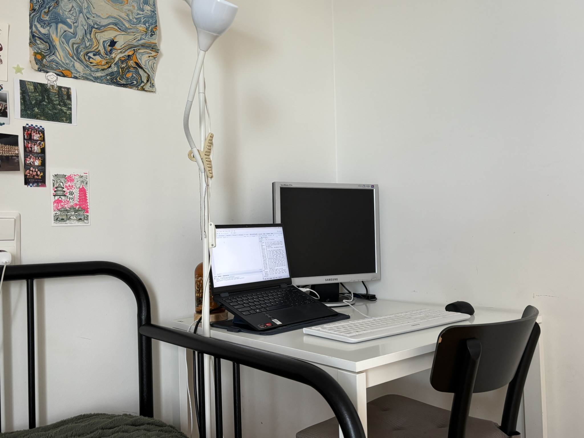 A table in the corner of the room squeezed between a bed and the wall. On the table there is a laptop, a monitor, keyboard and mouse which fills up majority of the tabletop space.