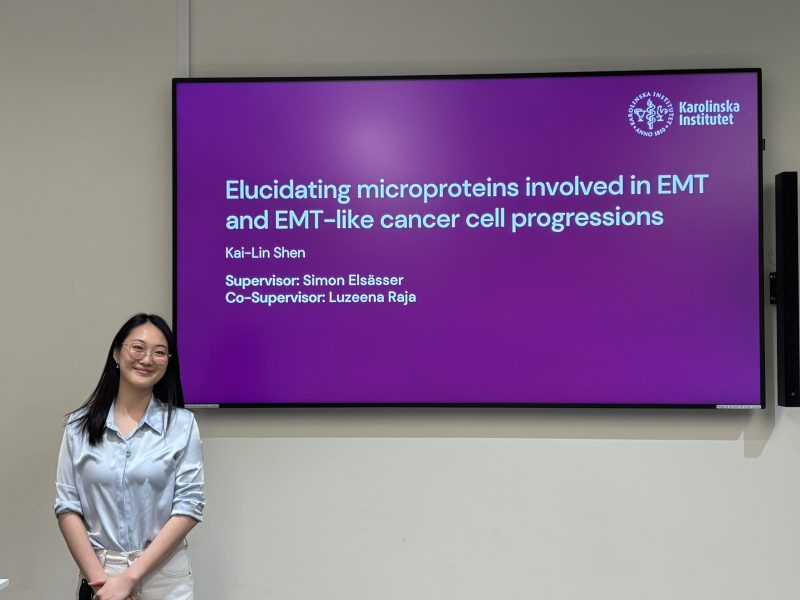 A girl stands in front of a TV screen displaying a powerpoint presentation of her Master's thesis title.