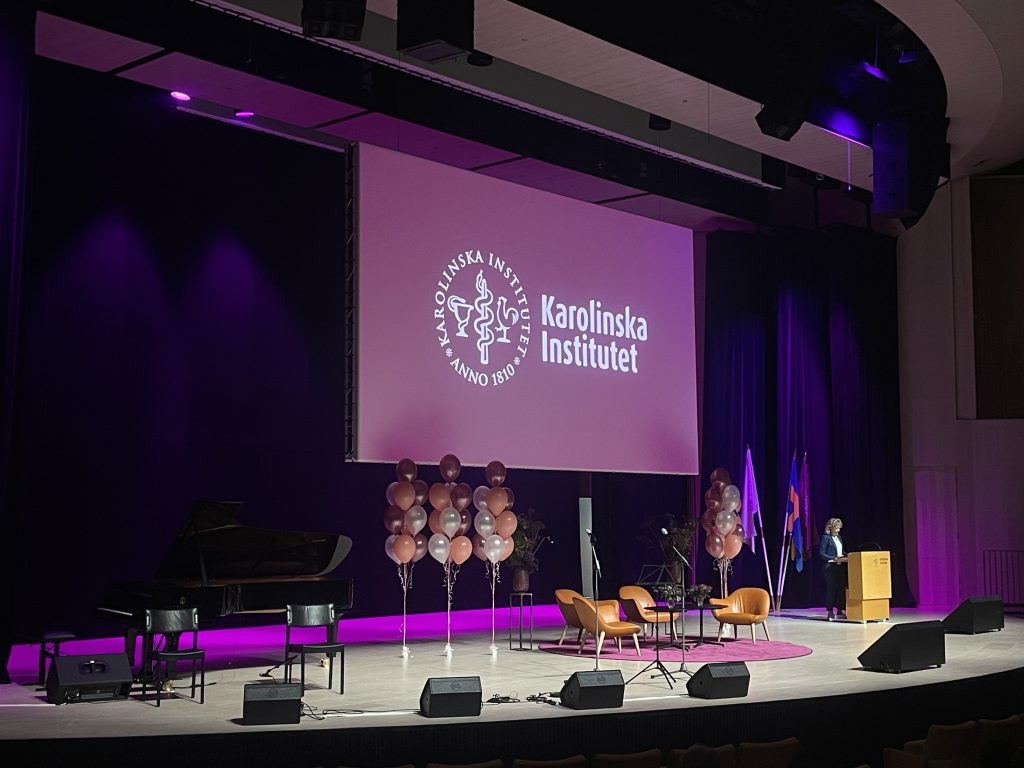 Stage decorated with globes, in the center some chairs and microphones, a piano on the right. In the back, a screen shows the logo of Karolinska Institutet.