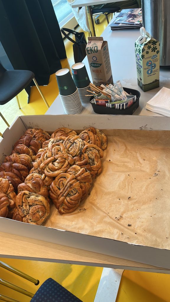 A box of Kardemummabullar, coffee, milk and paper cups on a table in a classroom.